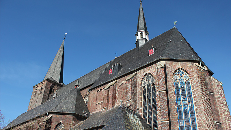 St. Maria Magdalena Kirche in Sonsbeck mit zwei hohen Türmen, rotem Backstein und großen Buntglasfenstern unter blauem Himmel