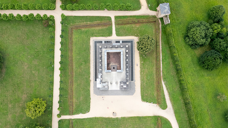 Luftaufnahme eines quadratischen, historischen Gebäudes umgeben von gepflegtem Gras und Wegen, mit Bäumen in der Nähe.