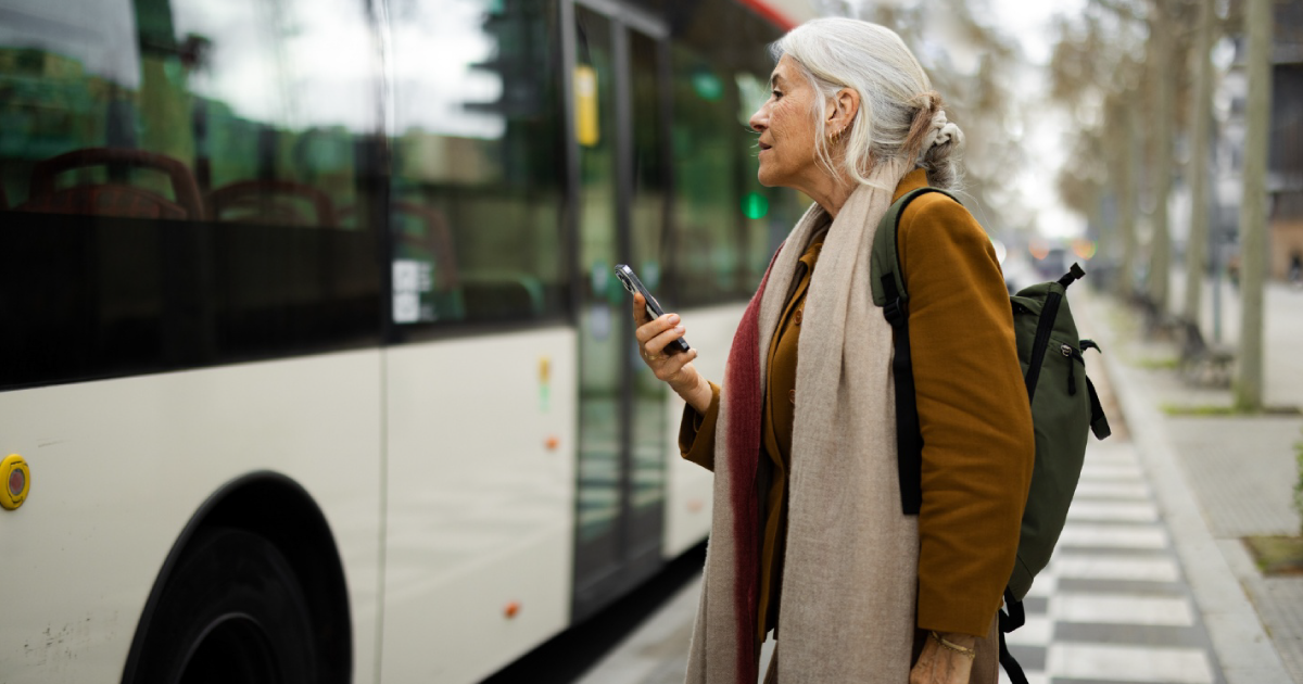 An older woman with a backpack holds a smartphone while waiting for a city bus at a sidewalk stop.