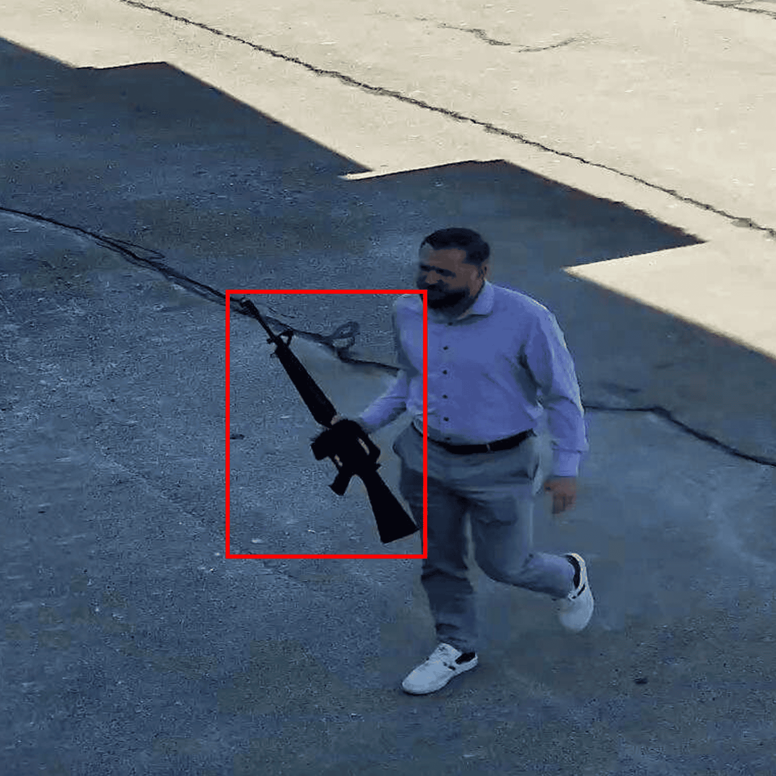 Man in light blue shirt and gray pants running on concrete ground while holding a black rifle.