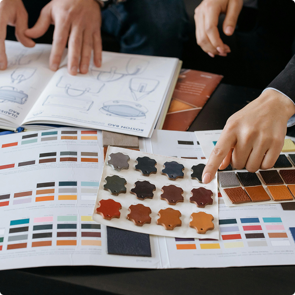 Two hands pointing at color swatches and sketches on a table, focusing on leather samples and design drawings.