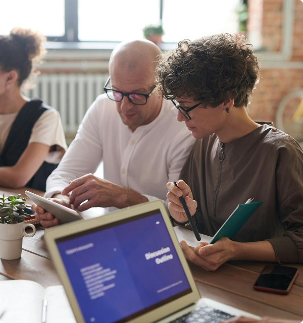 Two people wearing glasses collaborating over a tablet in a well-lit office with a laptop showing a discussion outline in the foreground.