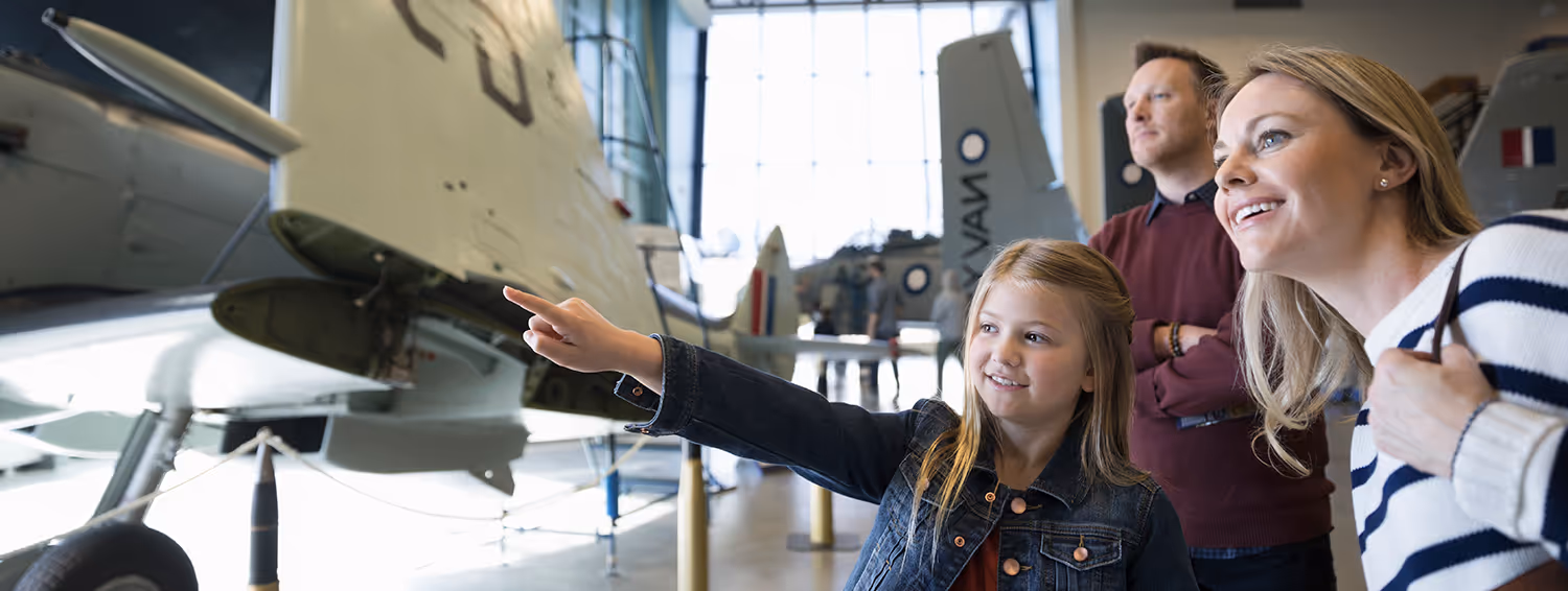 Young girl pointing at an aircraft inside a museum while her mother and father look on.