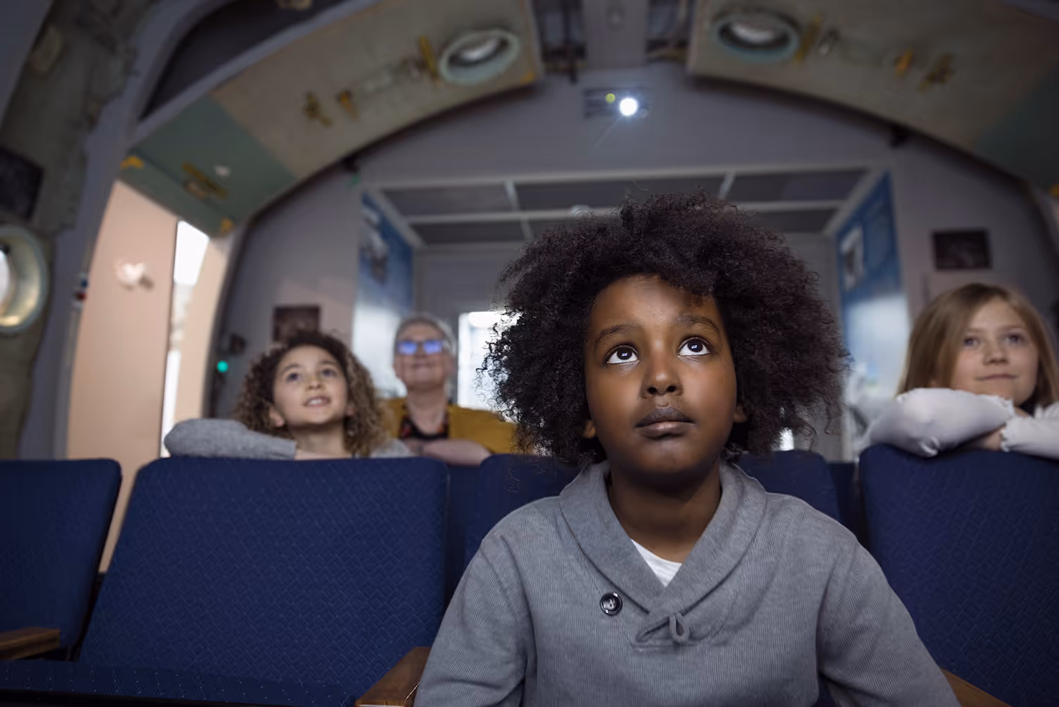 Four children and an adult seated in a theater or classroom looking intently forward, with the child in front having curly hair and wearing a gray sweater.