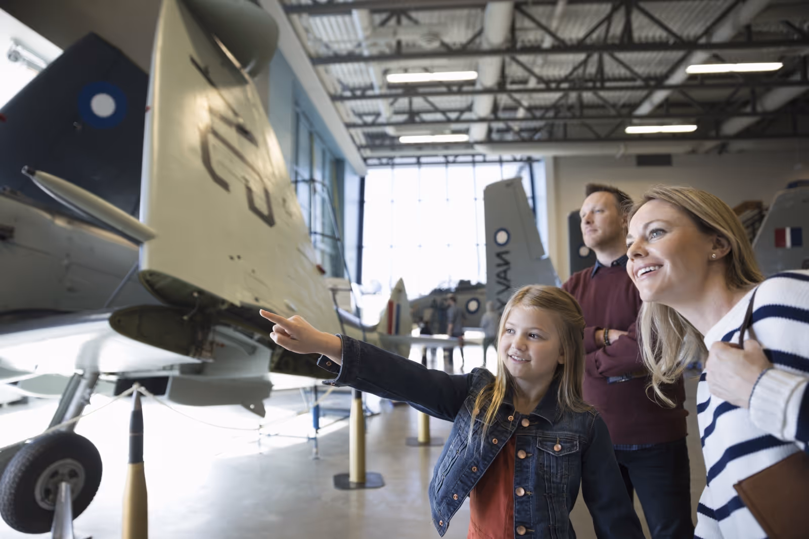 Young girl pointing at a vintage military aircraft inside an aviation museum, accompanied by a smiling woman and a man.