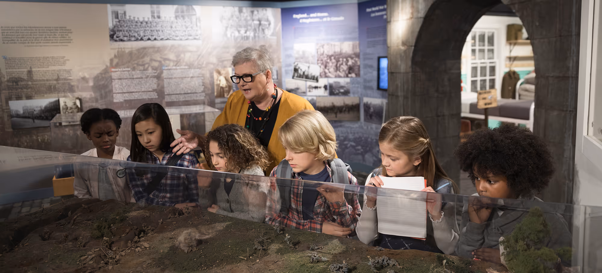 Teacher explaining a historical diorama to a diverse group of attentive schoolchildren in a museum exhibit.