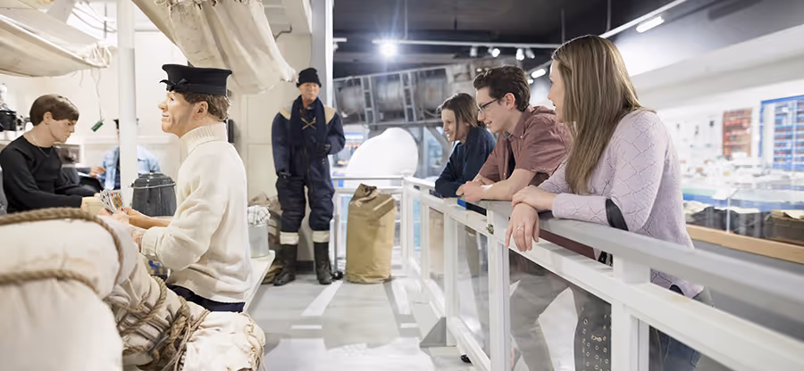 Three people observing maritime mannequins dressed in sailor uniforms on a ship exhibit.
