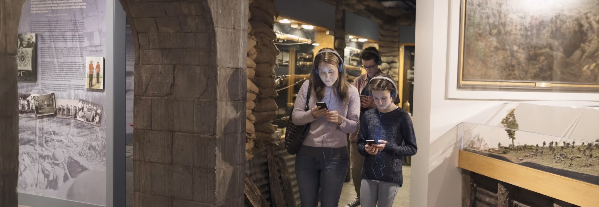 Three people wearing headphones and using smartphones while walking through a historical museum exhibit with sandbag walls and display cases.