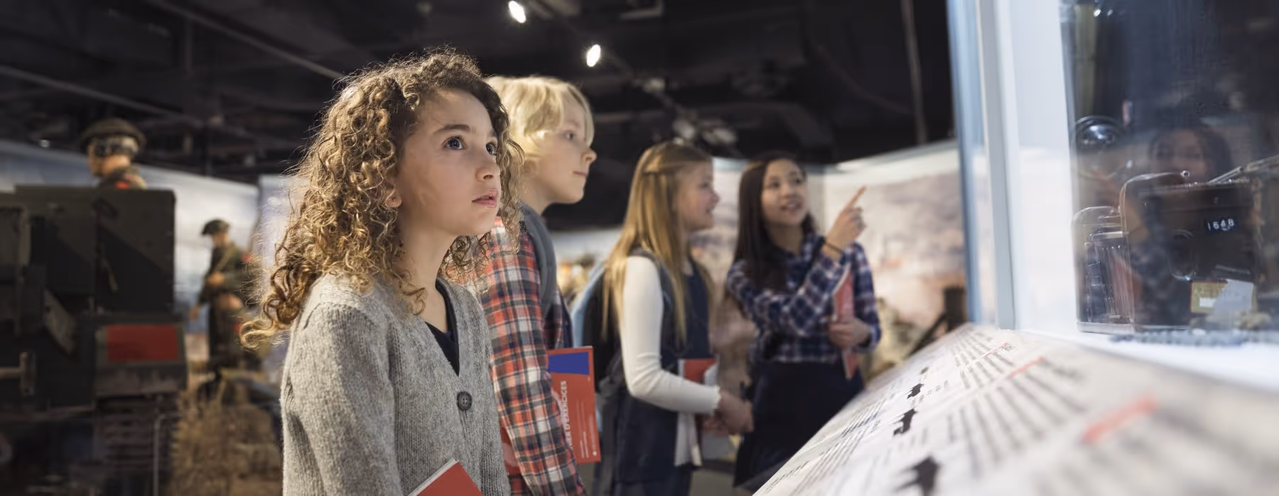Four children attentively looking at a museum exhibit behind glass, holding red booklets.