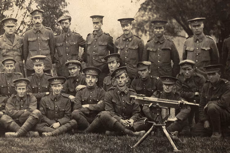 Black and white photo of 19 soldiers in uniform posing outdoors with a tripod-mounted machine gun in front.