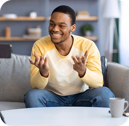 Smiling young man in a yellow and white shirt sitting on a couch and gesturing with his hands during a video call.