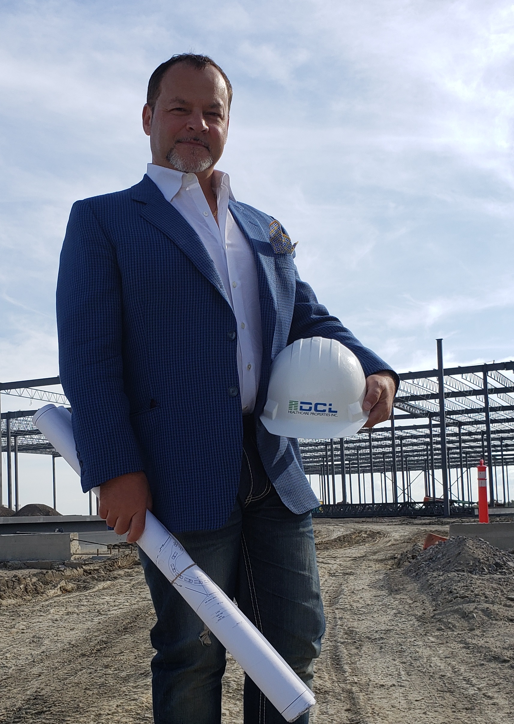 Man in a blue blazer holding a rolled blueprint and a white hard hat at a construction site with steel framework in the background.