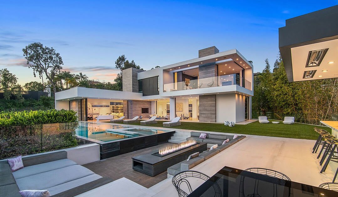 Modern two-story house with large glass windows, outdoor swimming pool, outdoor seating area with fire pit, and lawn at dusk.