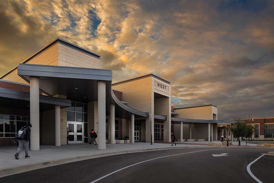 Modern school building entrance labeled 'WEST' with cloudy evening sky and students walking nearby.