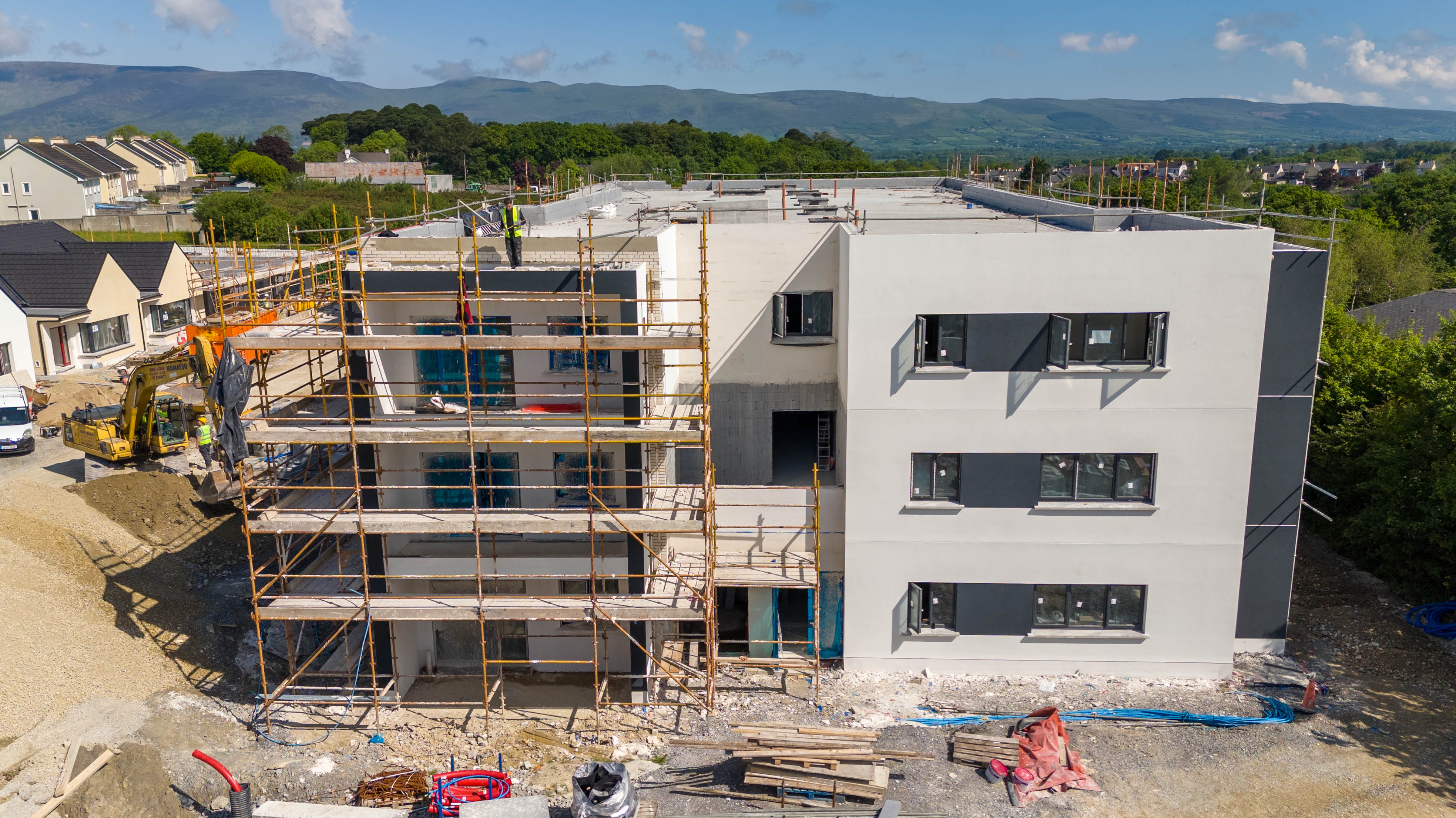Construction site of a modern multi-story building with scaffolding, workers, and machinery amidst a suburban landscape with houses and distant hills.