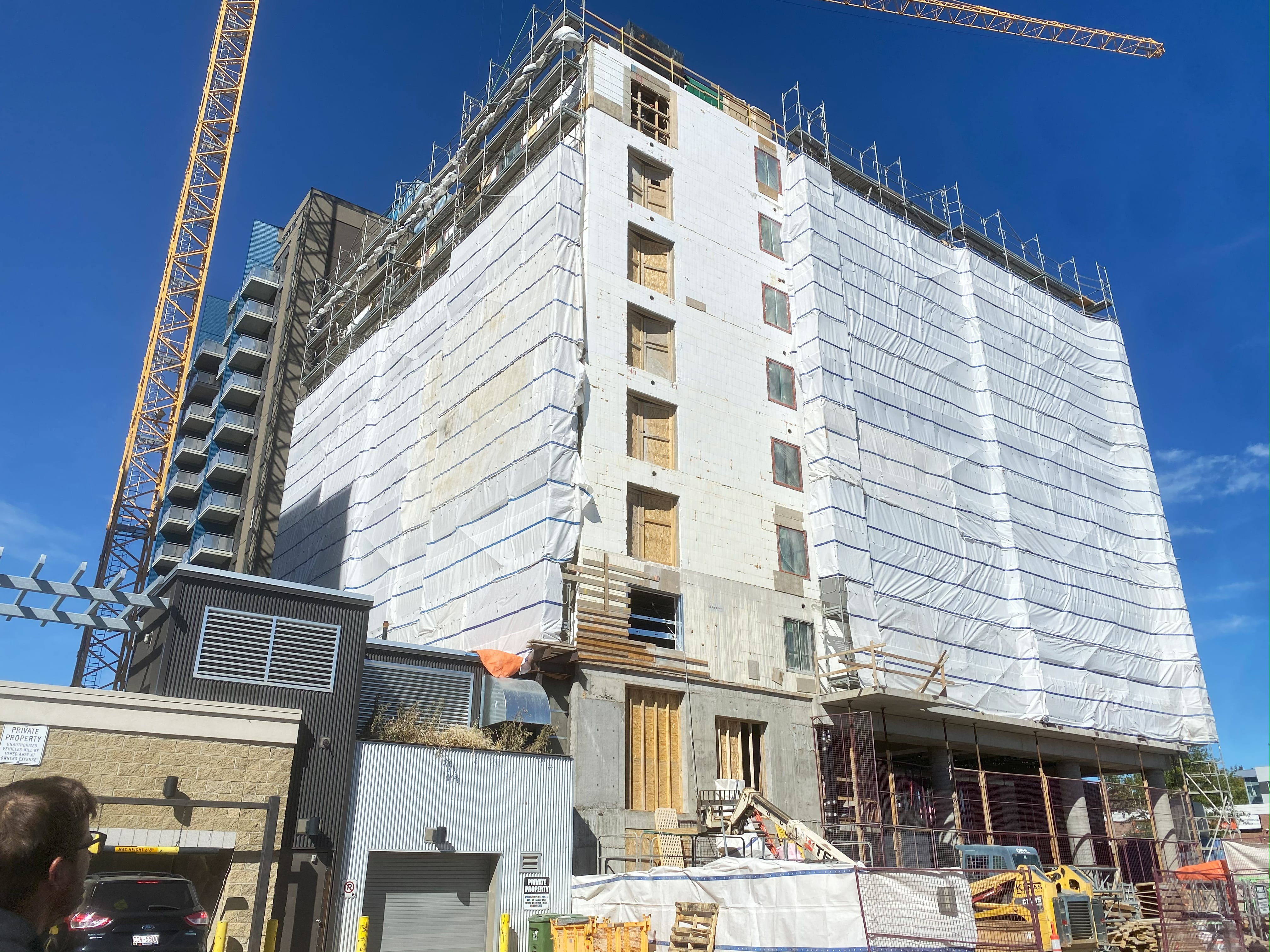 Multi-story building under construction covered in white protective sheeting with scaffolding and a yellow construction crane against a clear blue sky.