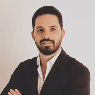 Homem de cabelos curtos e barba curta, vestindo camisa branca e blazer preto, posando com braços cruzados.