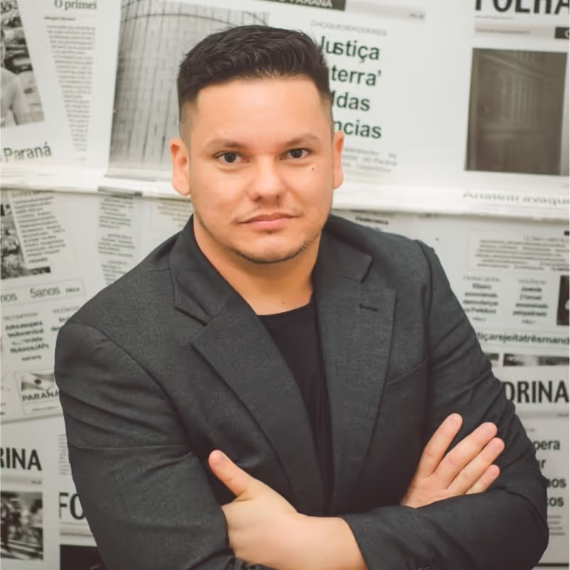 Homem com cabelo curto e escuro usando blazer cinza escuro, posando com os braços cruzados em frente a um fundo de jornais impressos.