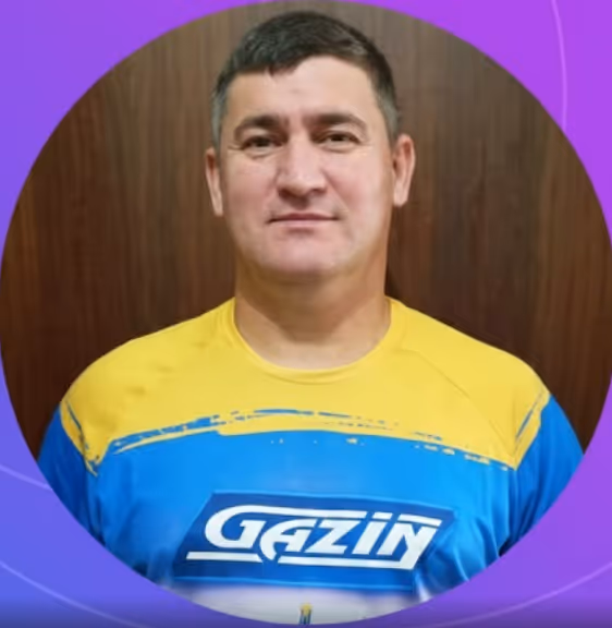 Homem com cabelo curto usando camiseta azul e amarela com a logo Gazin, fundo roxo e madeira.