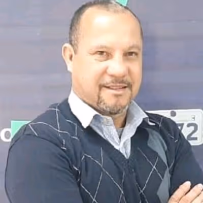Homem de meia-idade com cabelo curto e barba por fazer, vestindo camisa azul clara e suéter azul escuro com padrão xadrez, posando com braços cruzados.