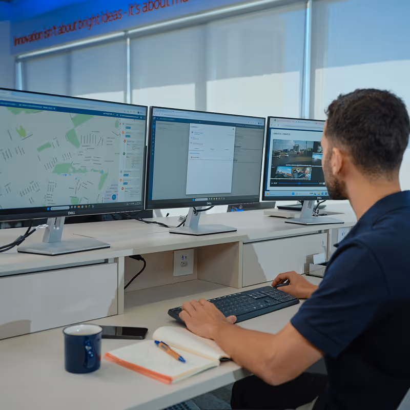 Homem sentado em uma mesa usando teclado e mouse, olhando para três monitores exibindo mapas e imagens de vigilância.
