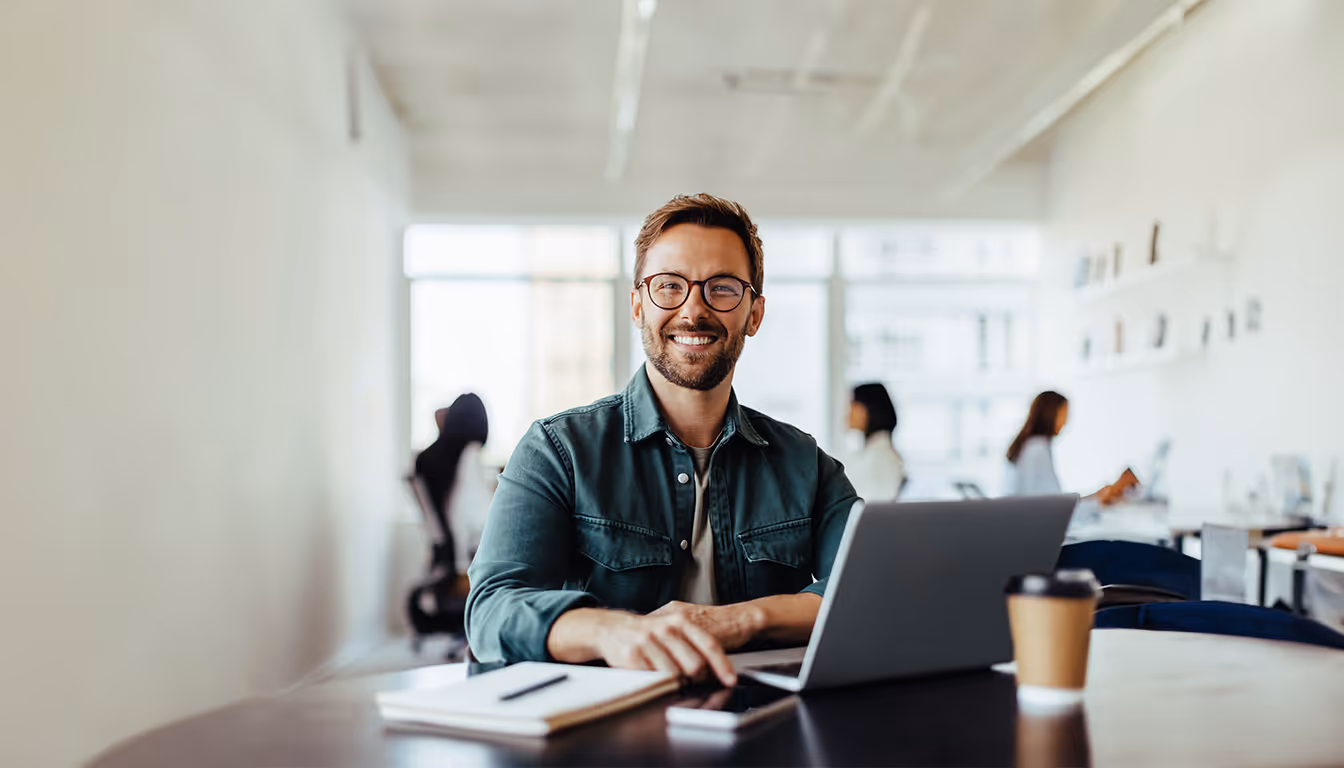 Homem sorridente usando óculos sentado em mesa com laptop, caderno e café em escritório moderno.