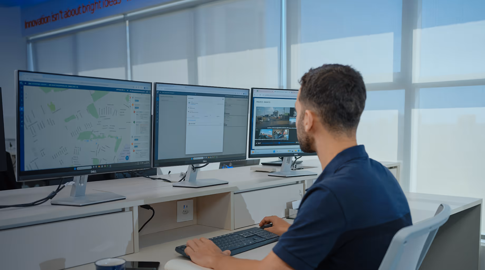 Homem trabalhando em escritório moderno com três monitores exibindo mapas, formulários e vídeos.