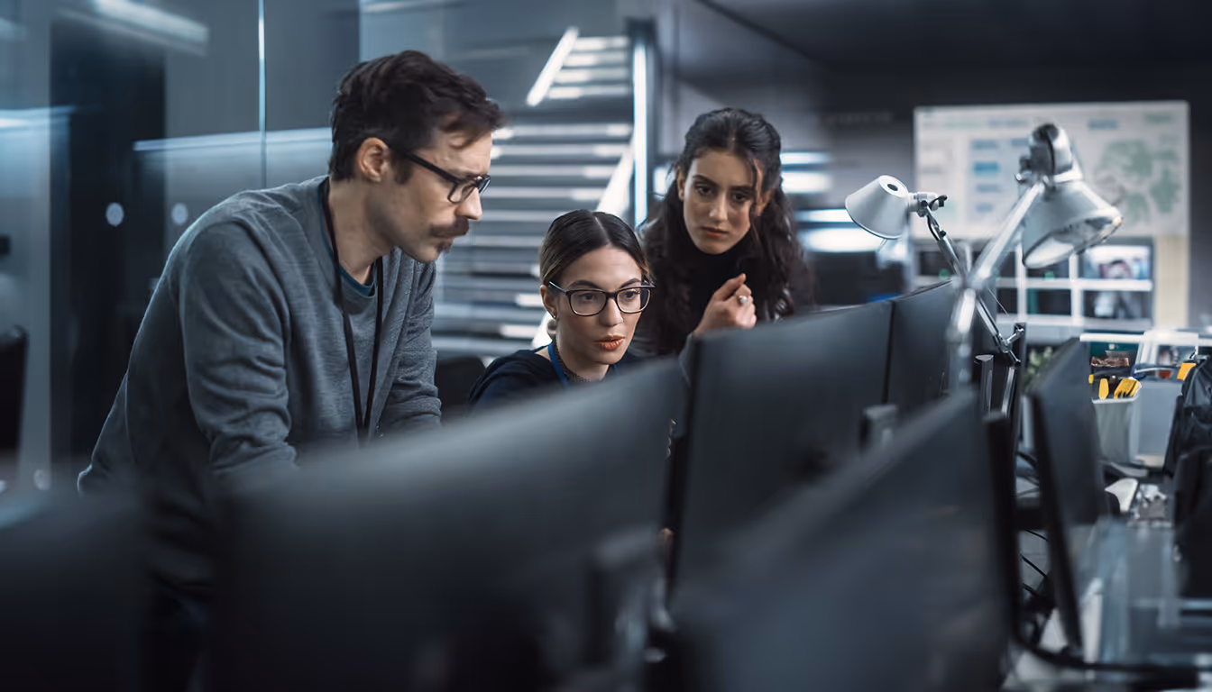 Três profissionais de tecnologia colaboram concentrados em frente a monitores em um ambiente de escritório moderno.