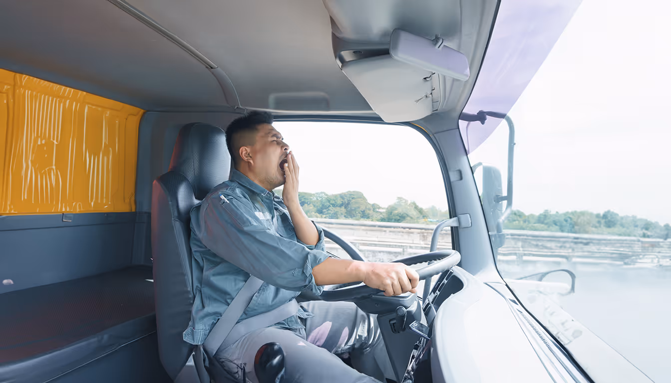 Motorista em um caminhão usando cinto de segurança, bocejando e aparentando cansaço ao dirigir.