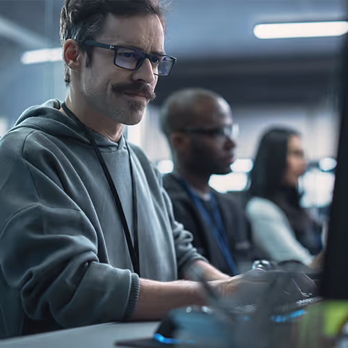 Homem de óculos e moletom cinza trabalhando concentrado em um computador, com duas pessoas desfocadas ao fundo.