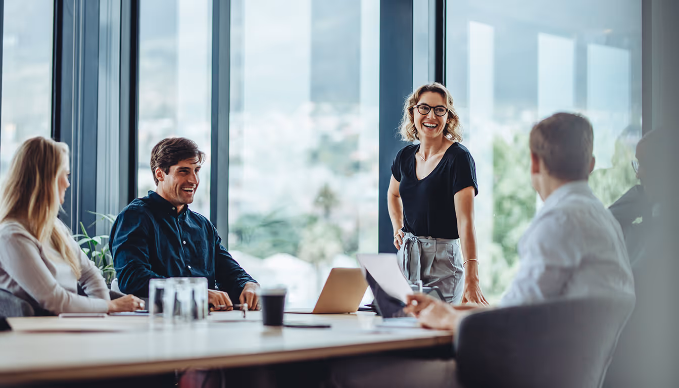Grupo de quatro pessoas sorrindo e conversando em uma reunião de negócios em uma sala com janelas grandes.