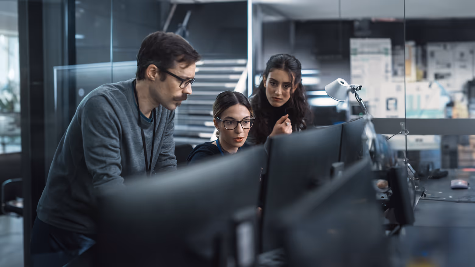 Três pessoas com expressão concentrada olhando para telas de computador em um ambiente de escritório moderno.