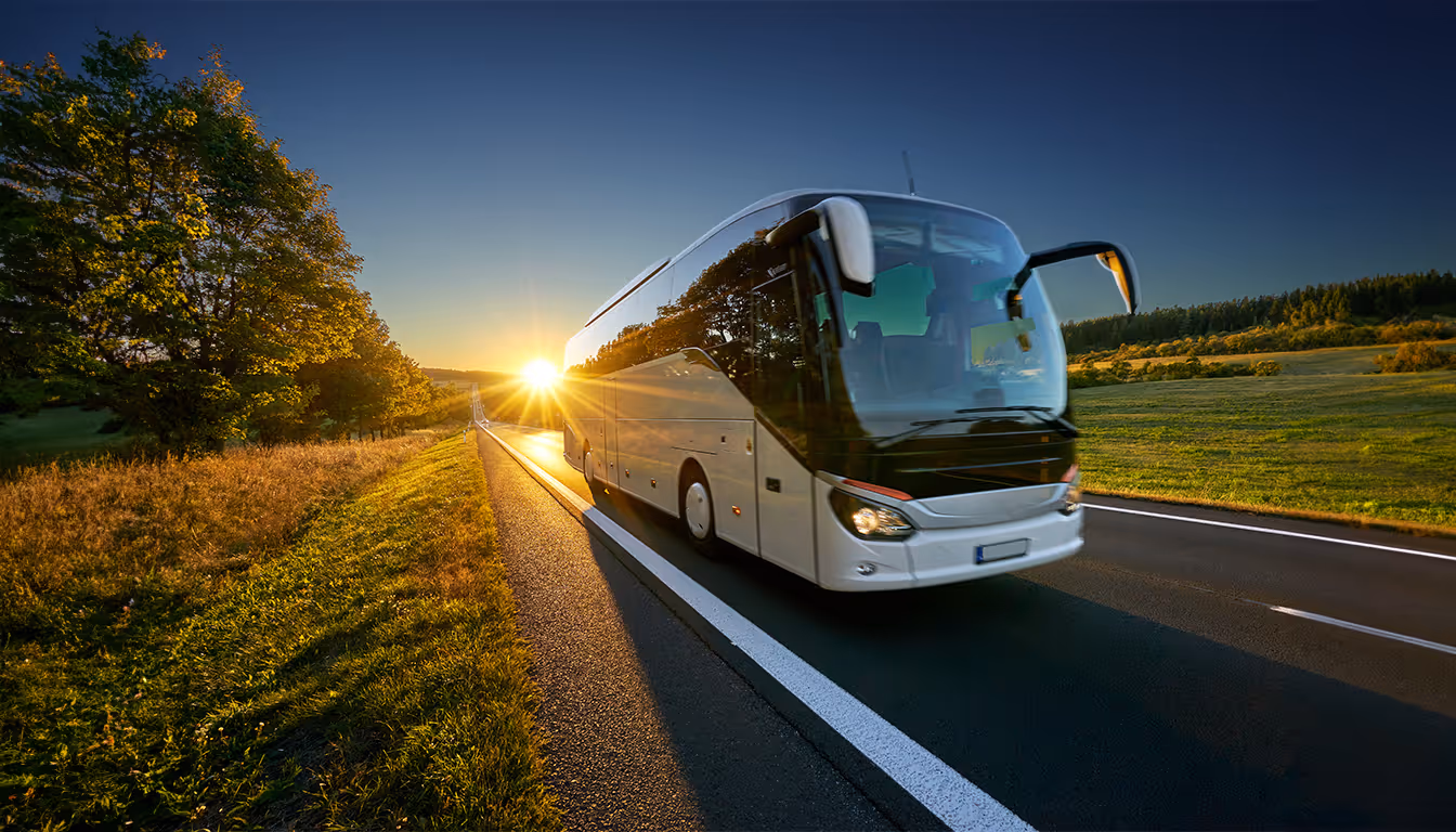 Ônibus branco moderno viajando em estrada rural ao pôr do sol com árvores e campos ao redor.