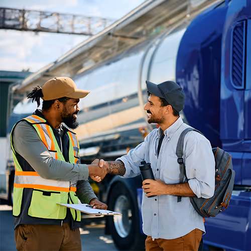 Dois homens apertando as mãos em um pátio de caminhões, um deles usando colete refletivo e boné, o outro com mochila e chapéu.