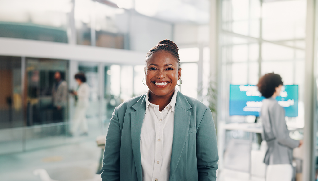 Pessoa sorridente vestindo blazer verde e camisa branca em um ambiente de escritório moderno e iluminado.