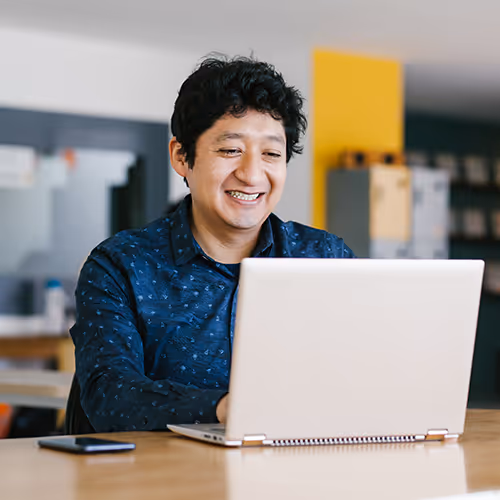Homem sorridente usando laptop em uma mesa de madeira em ambiente de escritório moderno.