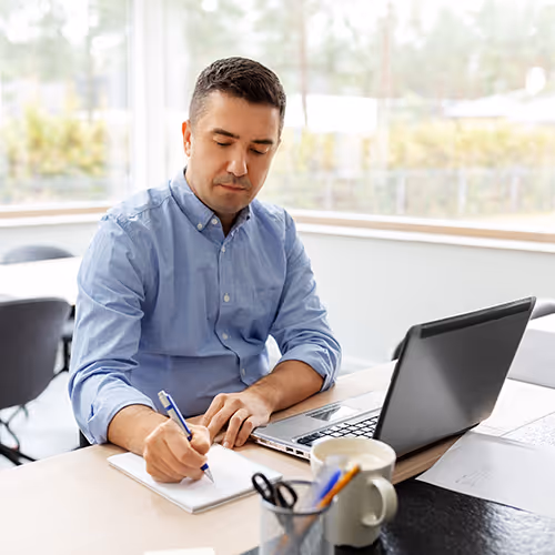 Homem sentado à mesa escrevendo em um caderno com laptop aberto ao lado em ambiente de escritório.