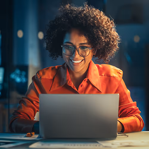 Mulher sorridente usando óculos e camisa vermelha sentada à mesa trabalhando em um laptop.
