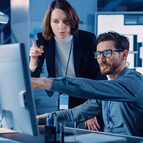 Homem sentado no computador apontando para a tela enquanto uma mulher observa e aponta, em um ambiente de escritório moderno.