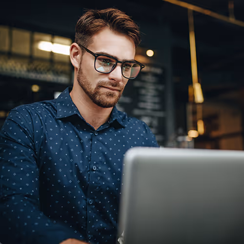 Homem usando óculos e camisa azul escura trabalhando em um laptop em ambiente interno com iluminação suave.