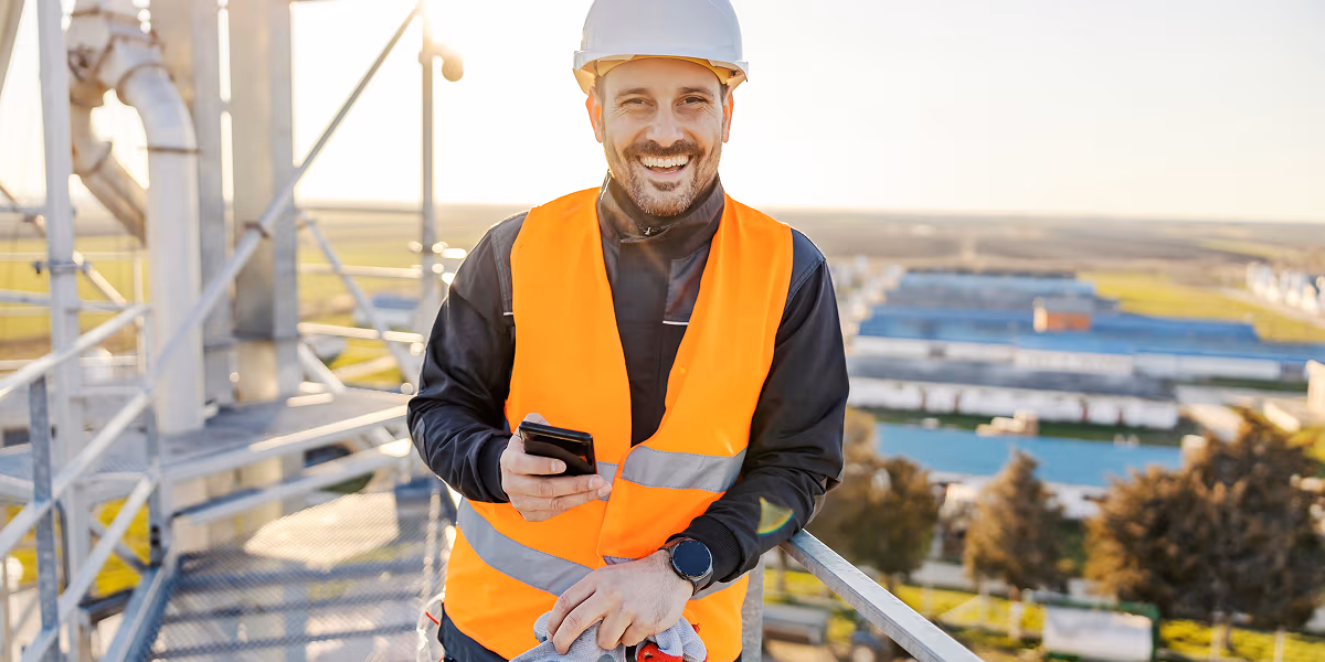 Smiling industrial worker wearing a safety vest and helmet stands on elevated platform, holding a smartphone—using WorkSafeAI’s voice-powered tool to instantly report a hazard or near-miss on-site, ensuring real-time safety compliance and data capture.