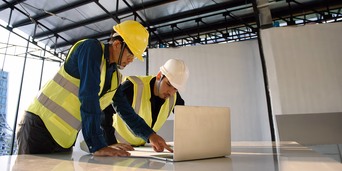 Two construction managers in safety vests and helmets review data on a laptop at a building site—evaluating real-time insights from WorkSafeAI’s voice-powered reporting system to understand incident trends and reduce operational costs through faster response and compliance.