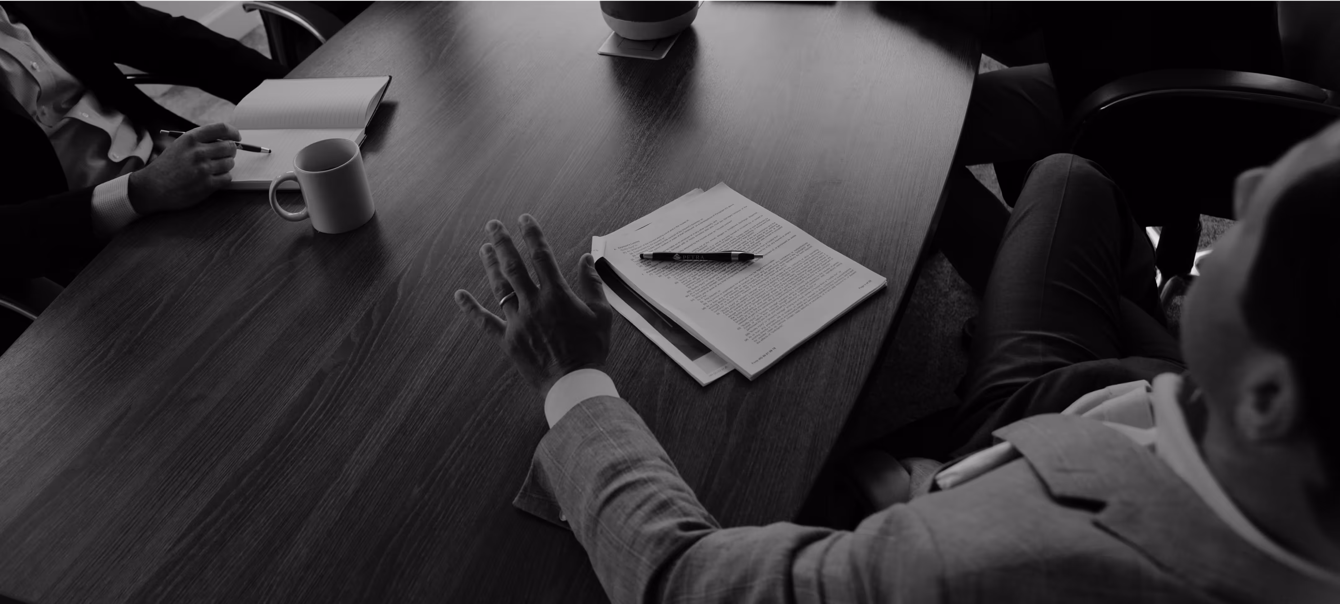 A black and white, high-angle photo of a business meeting in progress. One man gestures over a document on the table while another person takes notes.