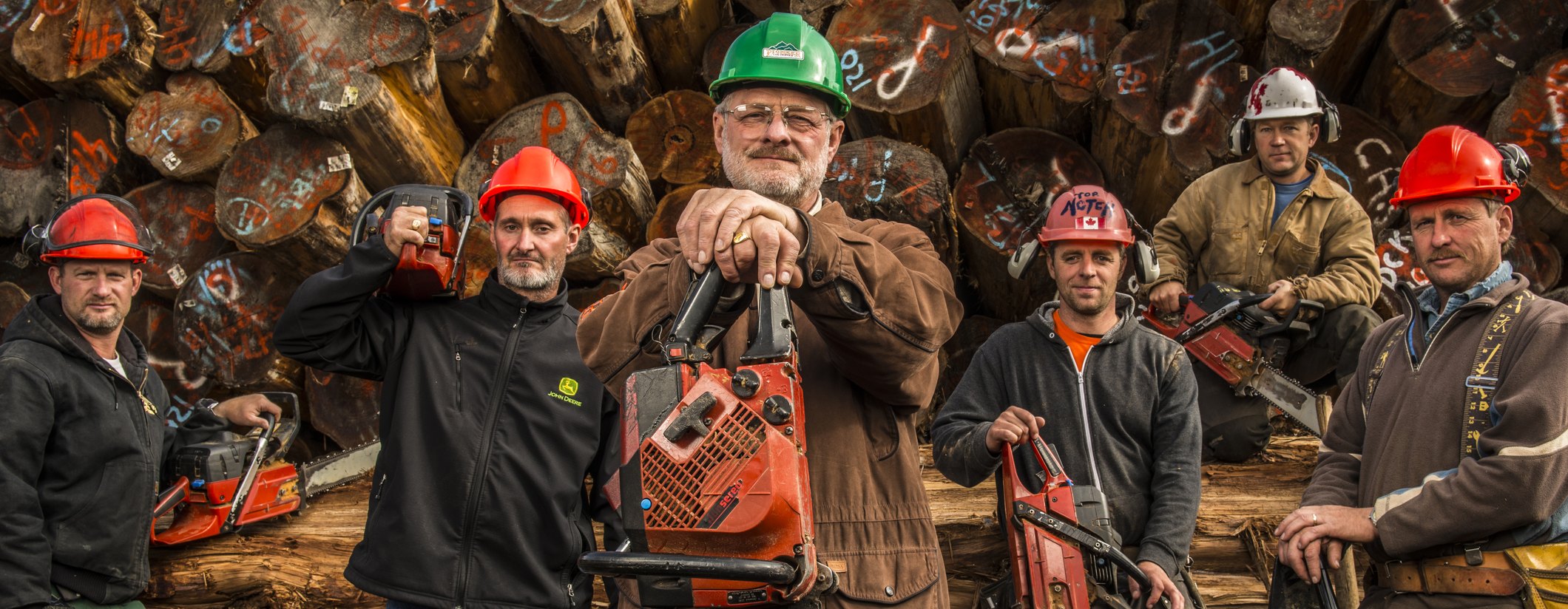 The Timber Kings crew holding chainsaws in front of a wall of logs
