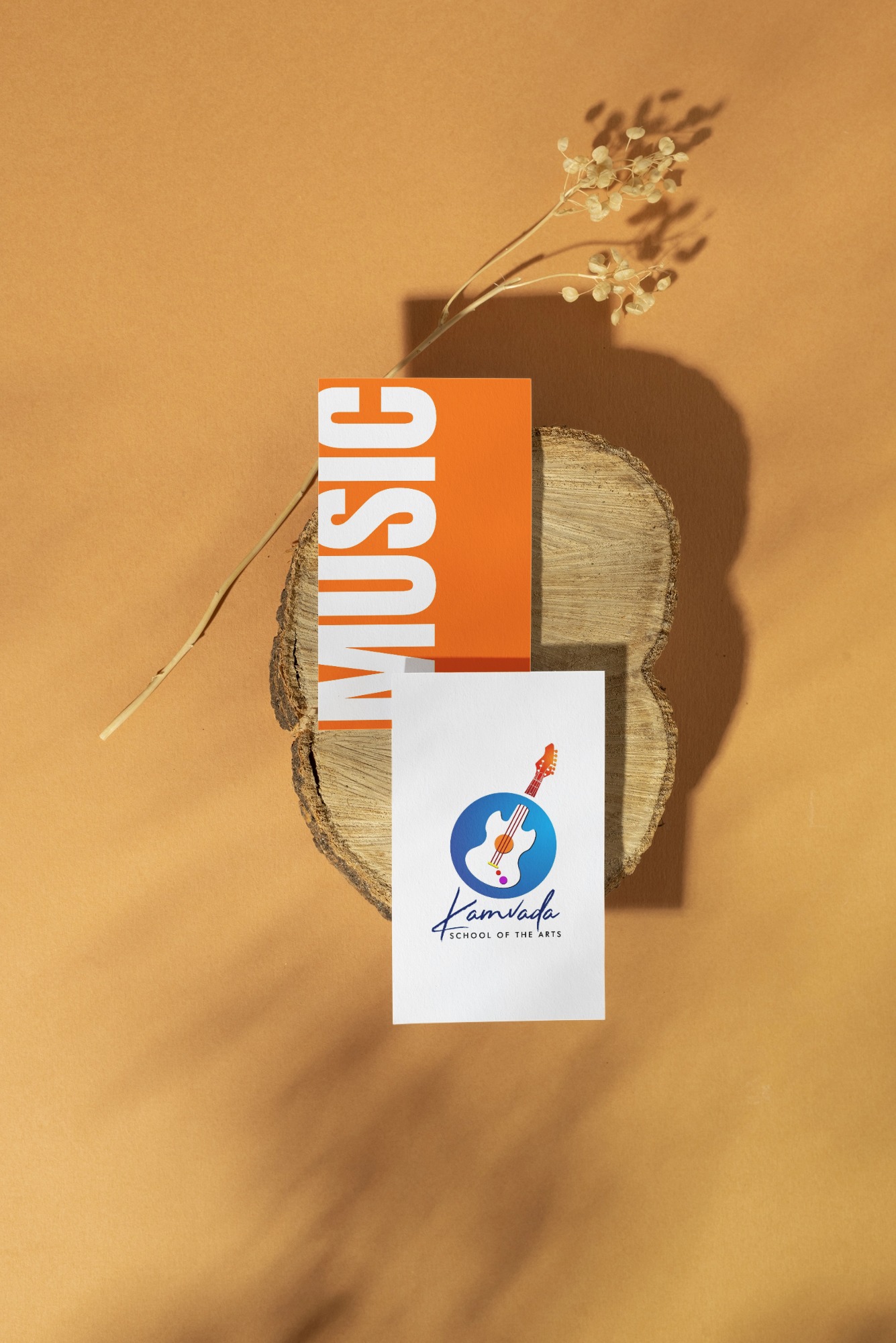 Two cards on a wood slice with dried flowers; one orange card with vertical text 'MUSIC' and one white card showing Kamvada School of the Arts logo featuring a guitar.