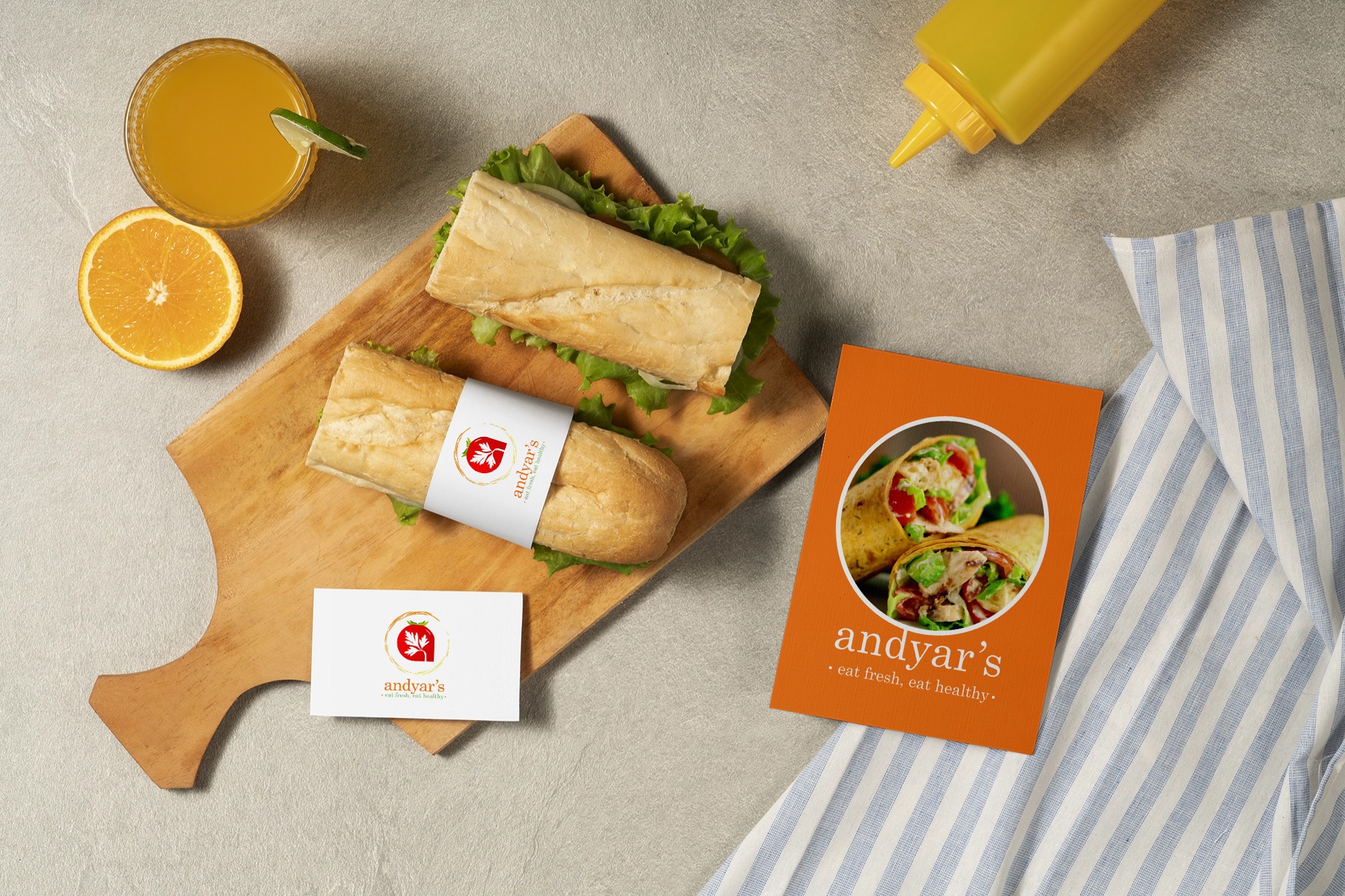 Two sandwiches with lettuce on a wooden board, an orange juice with lime slice, half an orange, a yellow mustard bottle, and Andyar's business card and menu on a gray surface with a striped cloth.