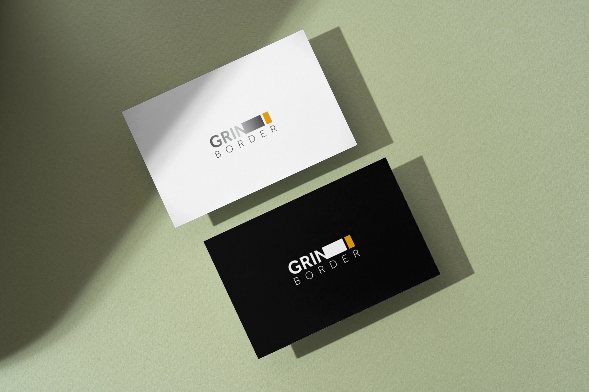 Two business cards on a green surface, one white and one black, both displaying the GRIN BORDER logo with a black and orange rectangular design.