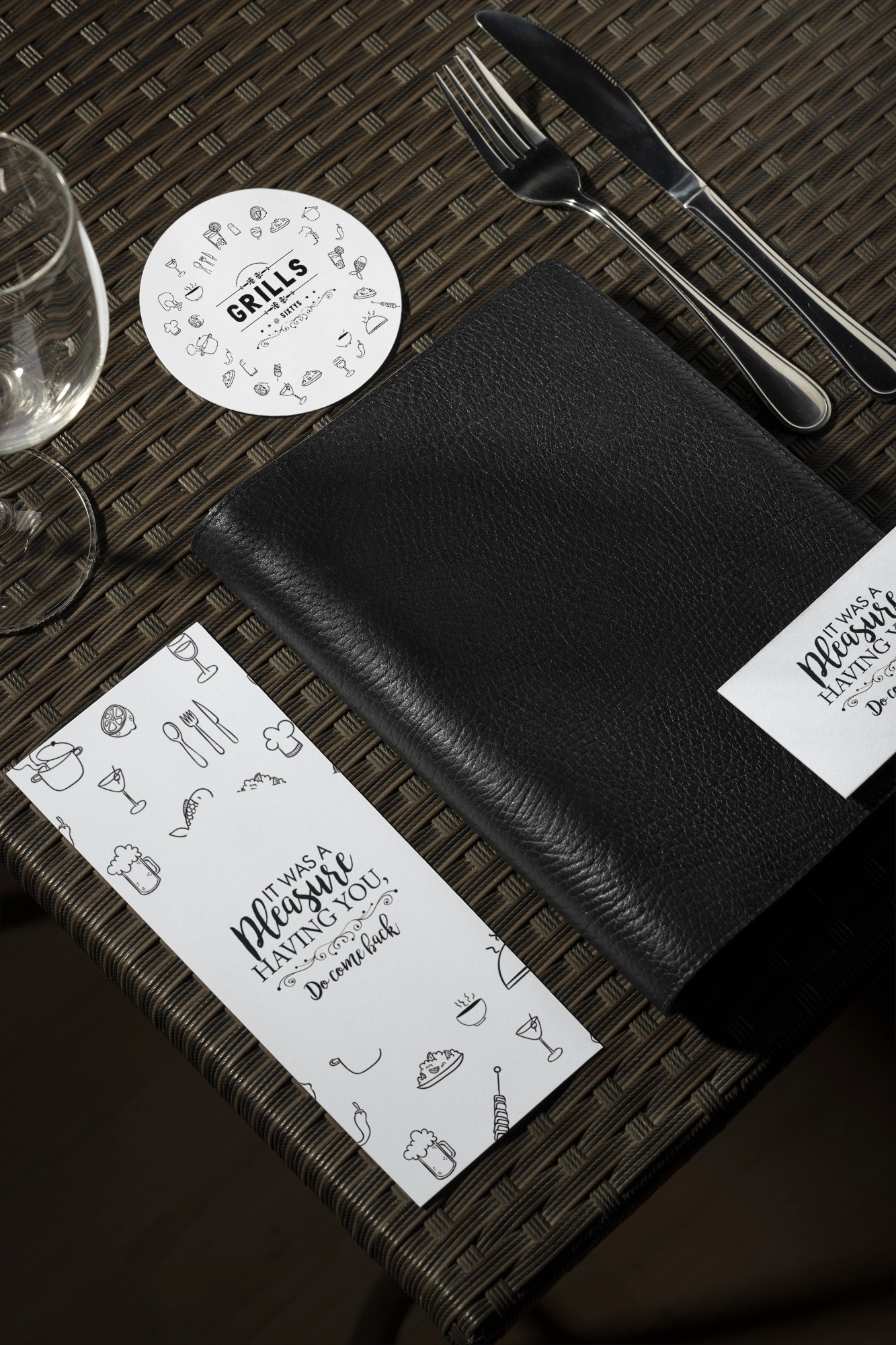 Table setting with fork, knife, empty wine glass, black leather menu, round coaster labeled GRILLS, and a card with the message 'It was a pleasure having you, Do come back.'