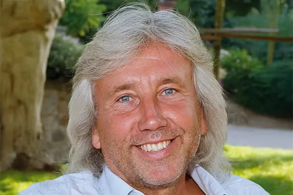 Mann mit schulterlangem grauem Haar und blauem Hemd lächelt draußen in einem Garten.