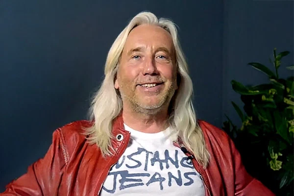 Christian Scheurer mit langen weißen Haaren trägt roten Lederjacke und weißes T-Shirt mit Aufschrift 'Mustang Jeans'.